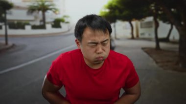 Şehir caddesinde kırmızı tişörtlü genç adam gün ışığında rahatsız edici bir ifade sergiliyor. Arka planda bulanık ağaçlar ve binalar var..