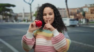 Kadın kalp şeklini korurken şehir caddesinde canlı bir süveter içinde hareketsiz duruyor oyuncu ifadeler sergiliyor ve açık hava manzarası sunuyor..