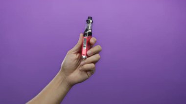 Mor duvar arkaplanına karşı Vape cihazı tutan adam, el ve cihaz tasarımını izole bir şekilde gösteriyor.