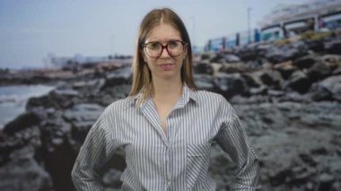 Sarışın, gözlüklü ve çizgili tişörtlü bir kadın kayalık sahil kenarında el ele tutuşuyor..