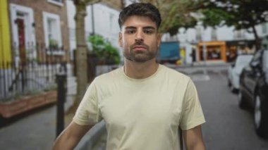 Hispanik adam yoğun şehir caddesinde yürürken kollarını esnetiyor. Gündüzleri binalarla dolu, kendine güveni tam..