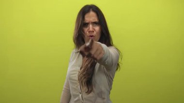 Kireç yeşili stüdyoda çatık kaşlı ve sert yüzlü bir kadın kamerayı işaret ediyor..