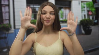 Sarı bluz giyen genç beyaz kız iki elini kameraya kaldırıyor. Kendine güveni tam..