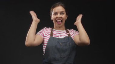 Avuç içleri görünen bir kadın, stüdyo önünde geniş bir gülümseme siyah bir arkaplan çizgili gömlek ve kot önlük giymiş; sürpriz bir kutlama.