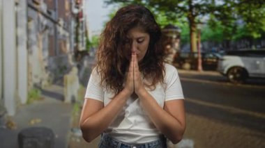 Beyaz tişörtlü genç İspanyol kadın ellerini kentin kaldırımlı bir caddesinde dua ederken; huzur yansıması duası.