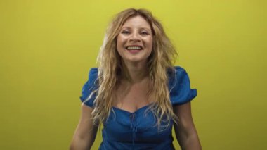 Avuç içlerini gösteren mavi bir elbise giyerken stüdyoda elini kaldıran ve arka planda gülümseyen bir kadın var..