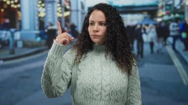 Kıvırcık saçlı, süveter giyen bir kadın, kalabalık şehir caddesinde kapalı şehir arka planında tasvip etmediğini belirterek baş parmak hareketi yapıyor..