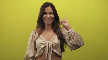 Yükseltilmiş işaret parmağı ve görünür eliyle gülümseyen İspanyol kadın stüdyoda göbeğini sarı duvarla gösteren dar bir bluz giyiyor; sevinç iyimserliği.