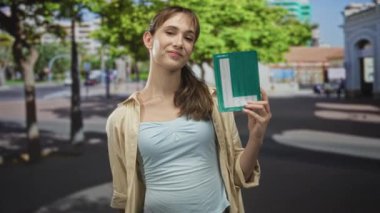 Elinde yeşil öğrenci işareti ve şehrin caddesinde ağaçların ve binaların yanında hafif bir gülümseme tutan kadın, gurur kaynağı..