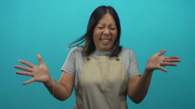 Turkuaz stüdyoda yüzünü kapatan bej önlüklü ve gri tişörtlü genç İspanyol kadın..