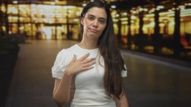 Bina girişinde eli göğsünde olan kadın, beyaz gömlek ve uzun saç görünür; minnettarlık samimiyeti.