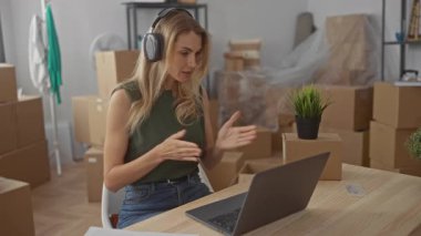 Yeni evde dizüstü bilgisayar kullanan bir kadın. Oturma odasındaki kutularla çevrili. Kulaklık takmış, günlük kıyafetler giyiyor..