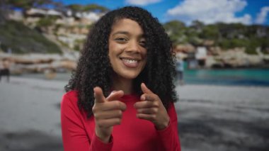 Kırmızı gömlekli kadın, kumlu sahilde açık mavi gökyüzünün altında iki işaret parmağını da kameraya doğrultuyor..