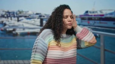 Renkli kazaklı genç İspanyol kadın güneşli bir günde arka planda teknelerle deniz kenarında jest yapıyor..