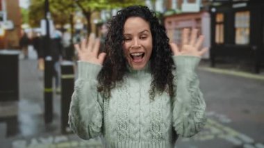 Açık havada, kıvırcık saçlı ve yeşil kazaklı bir kadın şehir arka planında canlı, bulanık bir şehir ortamında..