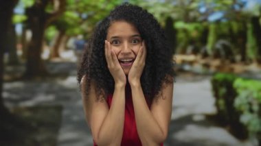 Yeşil bir parkta gülümseyen bir kadın, kırmızı bir gömlek giyiyor, kıvırcık saçları güneş ışığıyla aydınlatılıyor ve ağaçlarla çevrili, neşe ve huzur dolu bir anı yakalıyor..