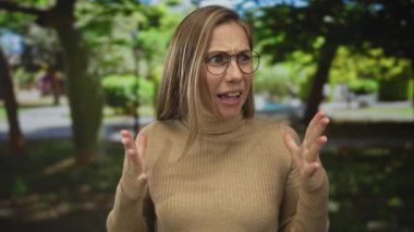 Parkta sürpriz yapan bir kadın, açık havada yeşil ağaçlar, kahverengi süveter ve gözlük takıyor, güneşli bir günde beklenmedik anlar yaşıyor..