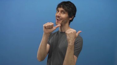 Koyu gri tişörtlü bir adam stüdyoda oyuncu bir sırıtışla iki başparmağını da işaret ediyor. Kendine güveni tam..