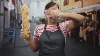 Bir baget tutan ve eldiven ile gözlerini kapatan bir kadın sokak kafe sahnesinde önlük ve çizgili bluz giyerek; neşeli promosyon.