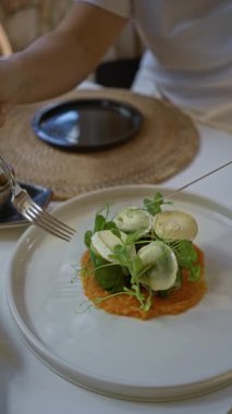 Akdeniz restoranında yemek yiyen bir kadın içeride gurme yemeğinin tadını çıkarıyor, taze yeşillikler ve zarif sofra takımı atmosferiyle karışık kaplamalar sergiliyor..