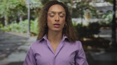 Açık ağızlı ve elleri yanaklarında olan bir kadın güneşli yeşil park yolunda, yapraklı gölgeliğin altında duruyor..