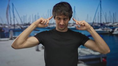 Liman iskelesinde elleri kulaklarıyla kulaklarını kapatan bir adam ve parlak gökyüzü altında demirlemiş tekneler;.