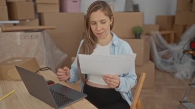 Hamile kadın yeni evinde açık dizüstü bilgisayarla dokümanları okuyor, paketlenmemiş kutularla çevrili, modern, aydınlık bir apartman dairesine taşınmanın heyecanını anlatıyor..