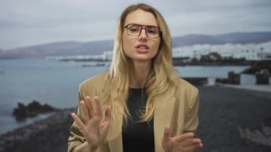 Elleri deniz kenarında sakince el sallayan, rüzgarda hafifçe savrulan sarı saçlı, gözlüklü ve kahverengi ceketli, gün ışığında sakin bir okyanus zeminine yaslanan bir kadın..