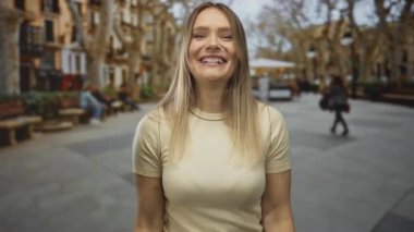 Genç sarışın kadın canlı bir şehir caddesinde mutlu bir şekilde gülümsüyor. Geçmişi bulanık, şehir ortamında enerji ve neşe saçıyor..