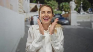 Açık şehir ortamında gülümseyen bir ifadeyle şehir caddesinde alkışlayan bir kadın.
