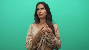 Kadın, elleri yeşil bir stüdyoda, göbek deliğini gösteren bej renkli bir bluz ve düşünceli bir yansıma ile parmaklarını birbirine gösteriyor..