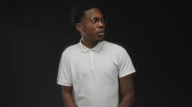 Gözlüklü ve beyaz polo tişörtlü adam karanlık stüdyo ortamında yana bakıyor; sessiz tefekkür.