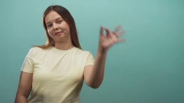 Sarı tişört giymiş, el işaretini göze yakın gösteren bir kadın, deniz mavisi stüdyosunda, oyuncu bir özgüven..