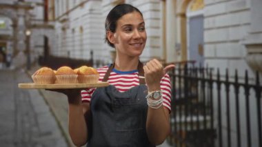 Sokakta elinde ahşap kek tepsisi tutan kadın, başparmağıyla önlük takarken pastaları işaret eden neşeli küçük işletme.