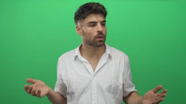 Beyaz gömlekli genç İspanyol adam, açık avuç içleri ve sorgulama ifadeleri olan yeşil bir stüdyoda ellerini açar..