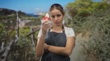 Eldivenli ve çapraz kollu çilekli dondurma bardağı tutan bir kadın yeşil ağaçların altında park yolunda kot önlük giyiyor..