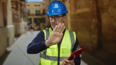 Güvenlik yeleği giyen ve kasketli bir adam, arka planda şehir sokağıyla dur işareti yapıyor.