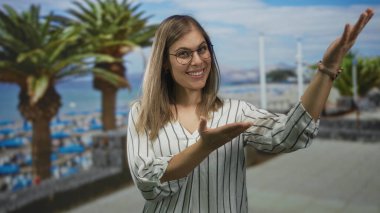 Sahilde palmiye ağaçlarıyla el kol hareketi yaparken gülümseyen gözlüklü bir kadın; mutluluk..