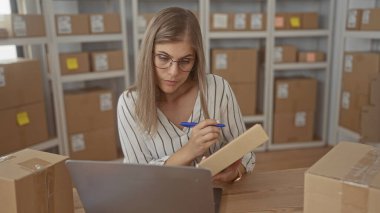 Sarışın kadın, ofis deposunda rafları olan dizüstü bilgisayarın yanındaki bir masada yazı yazıyor; verimlilik iş akışı verimliliği üretkenlik organizasyonu.