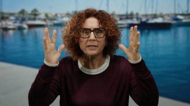 Sahilde duran bir kadın, çeşitli duygular ifade ediyor, arka planda tekneler ve su, açık havada sergilenen, orta yaşlı, esmer, İspanyol ve deniz manzarası.