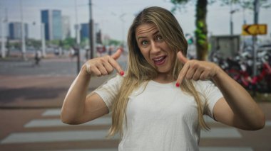 Gülümseyen sarışın kadın, yoğun güneşli şehir caddesinde yaya geçidini işaret ediyor..