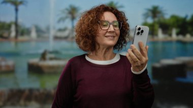 Güneşli bir parkta, palmiye ağaçları ve fıskiyeli bir telefon görüşmesinde elinde akıllı telefonuyla gülümseyen bir kadın.