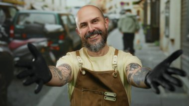 Dövmeleri ve eldivenleri olan kel sakallı bir adam. Şehir caddesinde sarılmayı teklif ederken gülümsüyor.
