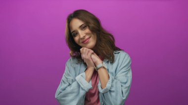 Pembe arka planda gülümseyen bir kadın, elleri sımsıkı bağlanmış, bir stüdyo ortamında neşe ve güven ifade eden, günlük mavi bir gömlek ve saat giyen, gençlik enerjisi sergileyen.