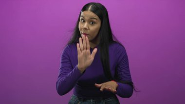 Kameraya bakan genç Latin kadın ellerini uzatarak el kol hareketleri yapıyor ve mor stüdyoda hafifçe başını eğiyor..