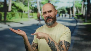 Şehir caddesinde sakallı, dövmeli, sarı gömlekli, açık havada güneş ışığı altında duran kel bir adam..