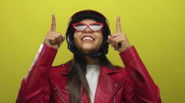 Kırmızı ceketli ve kasklı gülümseyen genç bir kadın izole edilmiş sarı bir arka plana iki eliyle işaret ediyor..