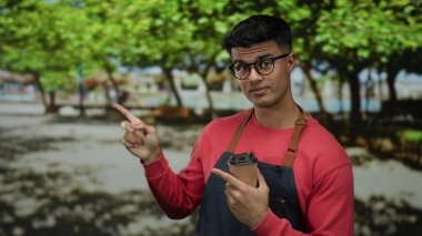 Parkta gülümseyen yakışıklı adam elinde kahve, kırmızı gömlek ve gözlüklerle açık havayı gösteren yeşil ağaçlarla çevrili parlak güneşli bir günü yansıtıyor..