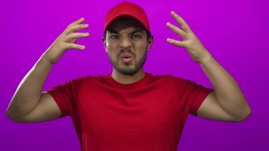 Kırmızı başlık ve kırmızı gömlek giyen kurye mor arka plana karşı stüdyoda iki eliyle gözlerini kapatıyor..