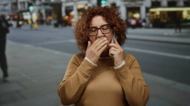 Orta yaşlı, esmer saçlı bir kadın kalabalık bir şehir sokağında telefonla konuşuyor, hareketli bir şehir geçmişinin ortasında jestler yapıyor..